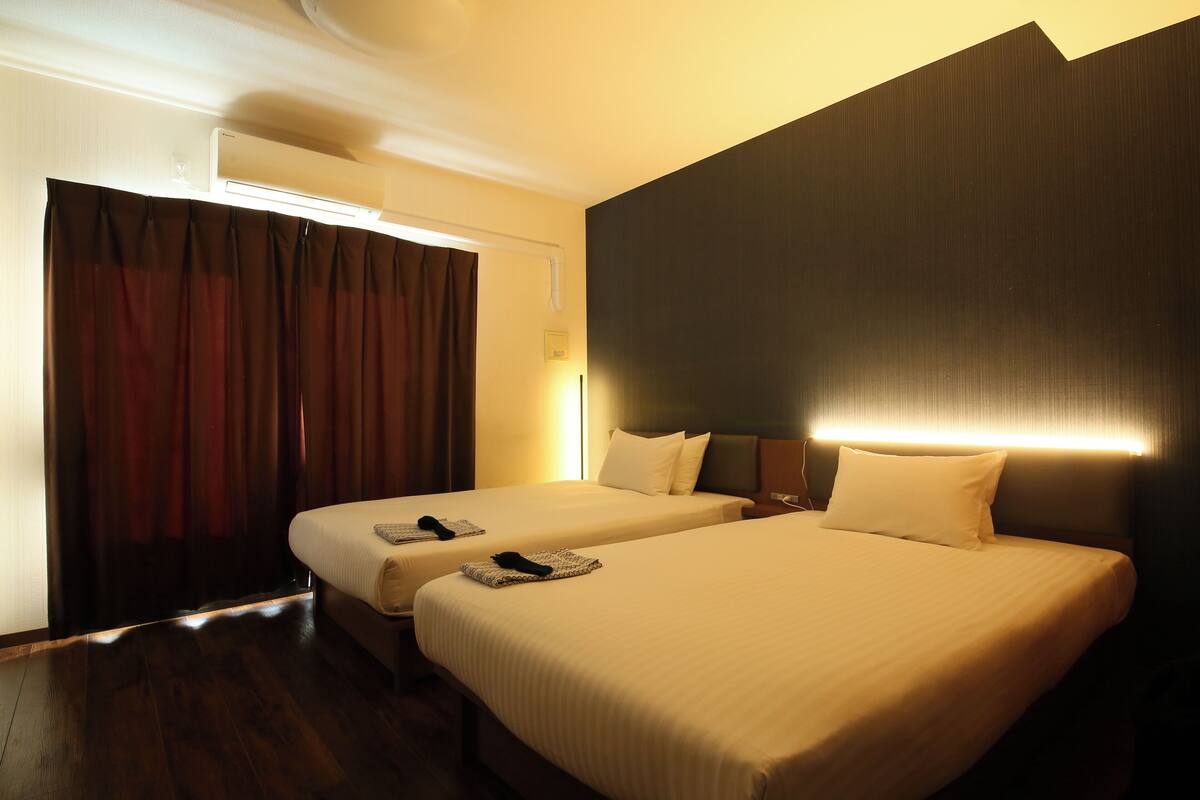 Une chambre d’hôtel avec deux lits, un mur sombre et une fenêtre avec des rideaux.