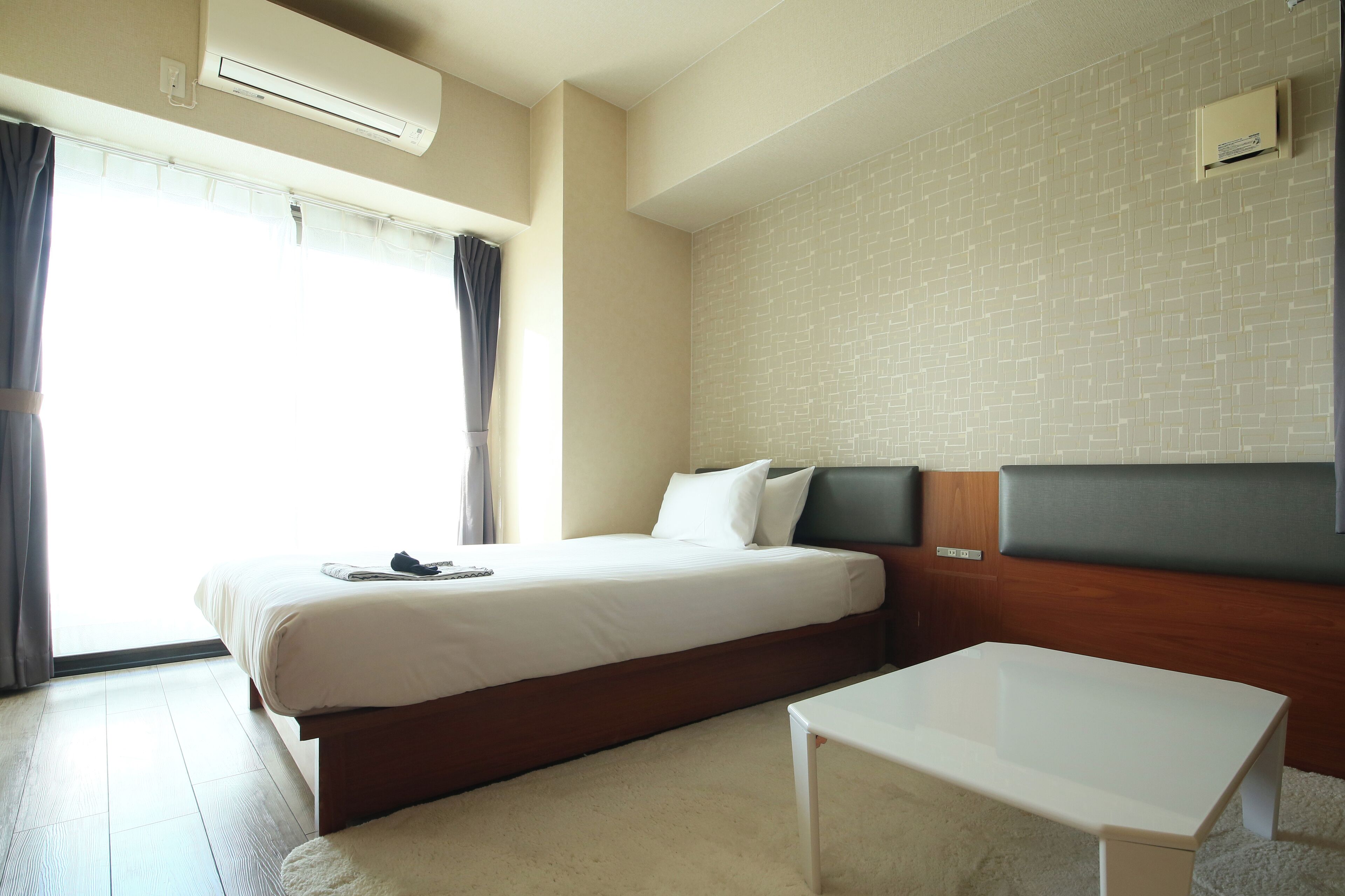 A hotel room with a large bed, a bedside table, a window with curtains, and a wall-mounted air conditioner.