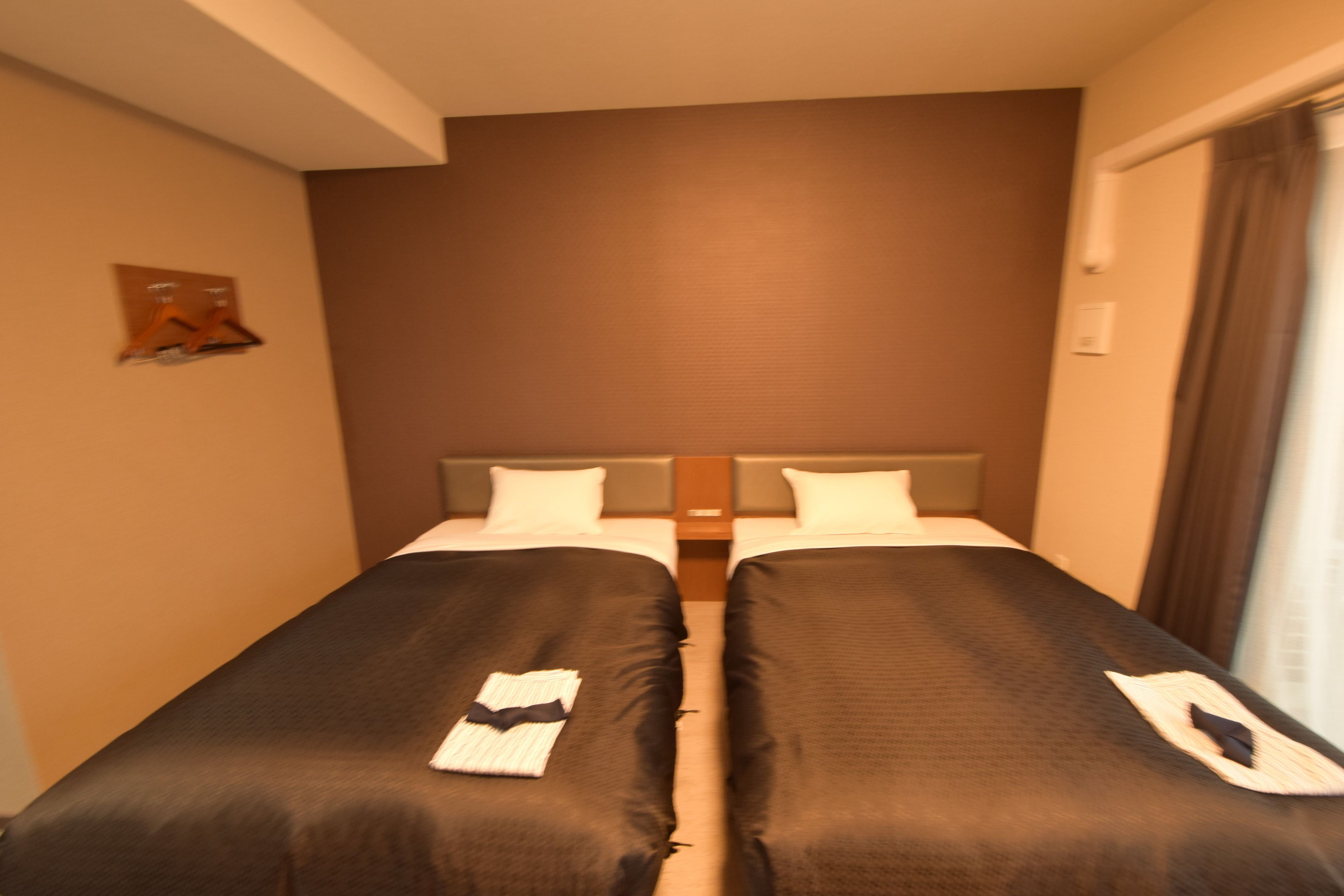 A hotel room with two single beds, a wooden headboard, a small wall-mounted shelf, and a window with curtains.