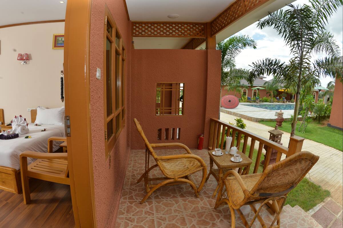 Habitación de hotel con dos camas, un balcón con mobiliario de mimbre y vistas a una piscina y follaje.