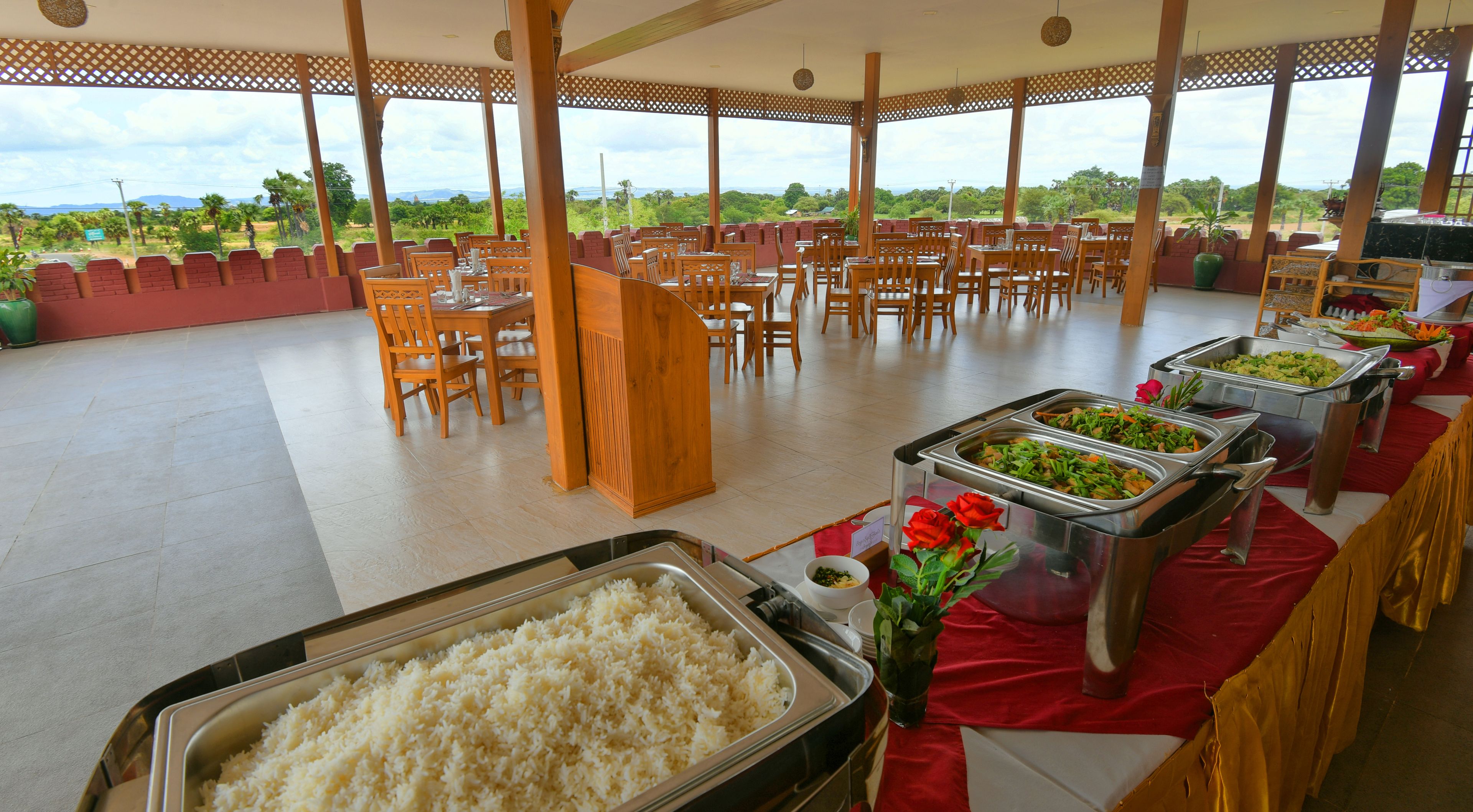 A buffet setup with multiple food stations, including rice and vegetables, in a spacious outdoor dining area with a view of greenery and distant mountains.