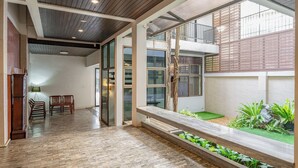 Amplo espaço interno com piso de madeira, bancos e vista para um jardim através de grandes janelas.