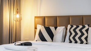 Uma cama bem arrumada com cabeceira, duas almofadas com padrão em ziguezague, luminária de mesa e uma tomada na parede.