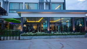 A modern hotel with a glass facade, outdoor seating area, and a green umbrella. - UDTEL Boutique Hotel (Udon Thani)
