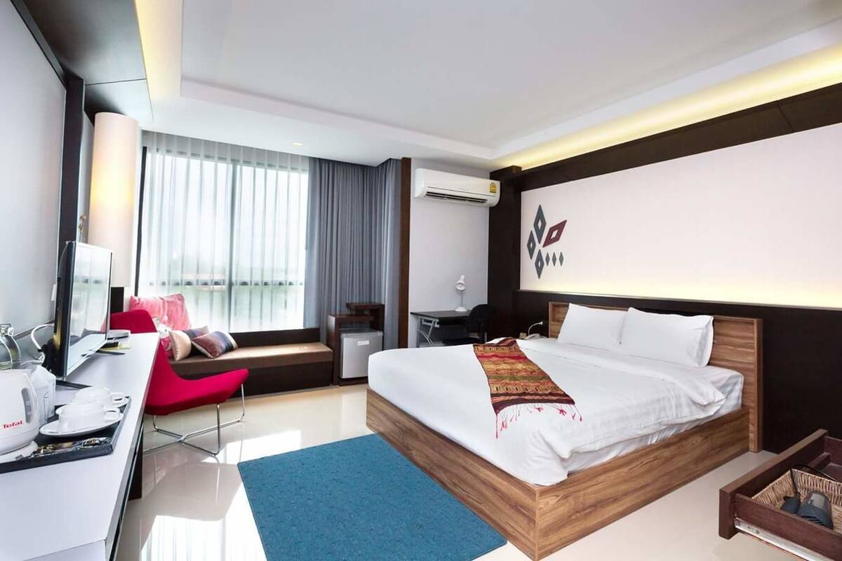 A modern hotel room with a large bed, a desk with a computer, a chair, and a small table with a coffee maker.