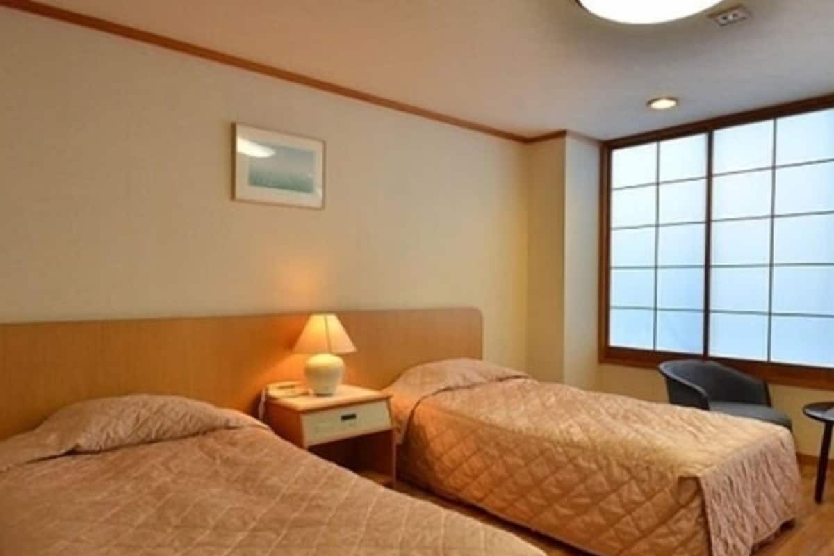 A hotel room with two beds, a nightstand with a lamp, a chair, and a window with blinds.