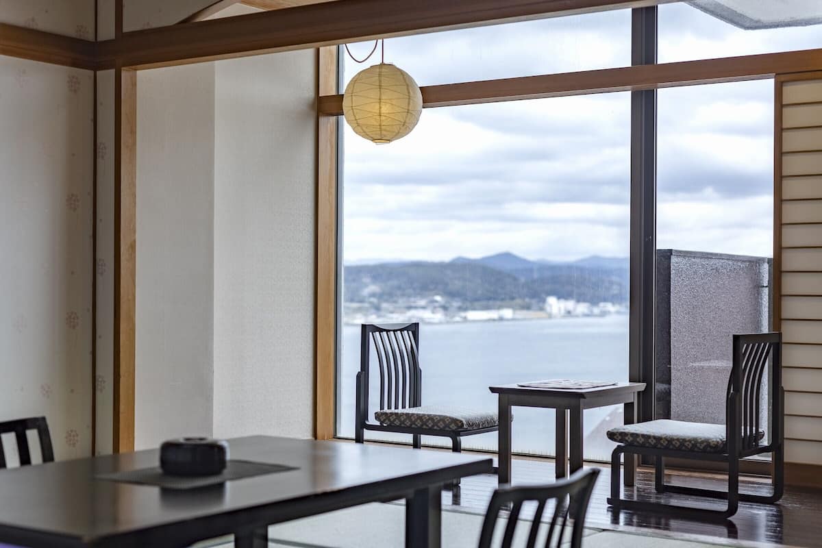 A room with a view of a body of water, a table, chairs, and a hanging paper lantern.