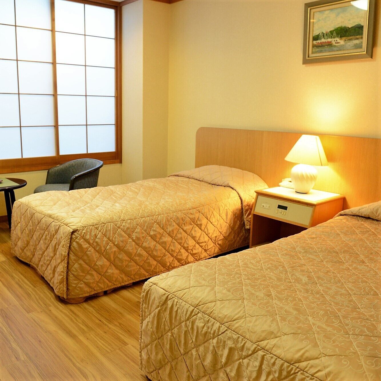A hotel room with two beds, a chair, a small table, and a nightstand with a lamp.