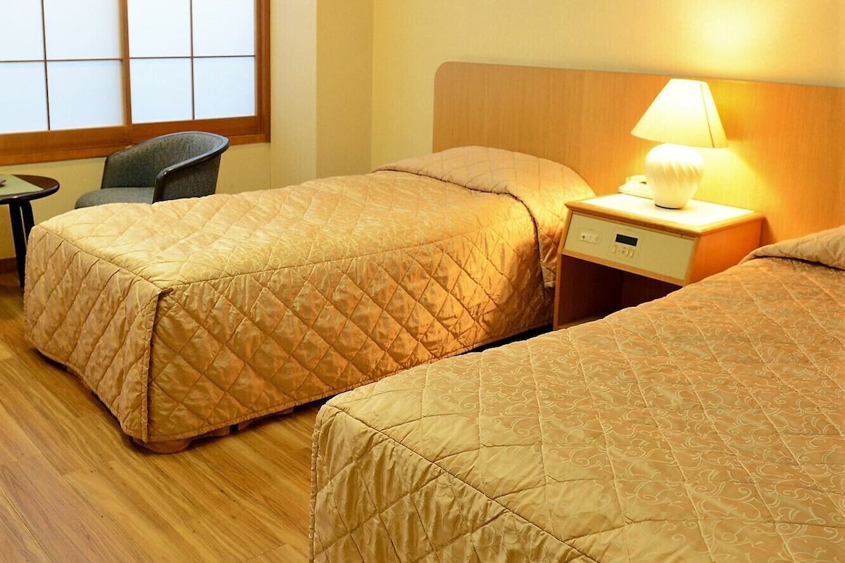 A hotel room with two beds, a chair, a small table, and a nightstand with a lamp.