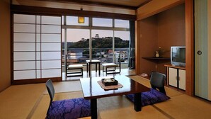 Una habitación japonesa tradicional con suelo de tatami, una mesa baja con cojines, un televisor en un mueble y vistas a un castillo a través del agua.