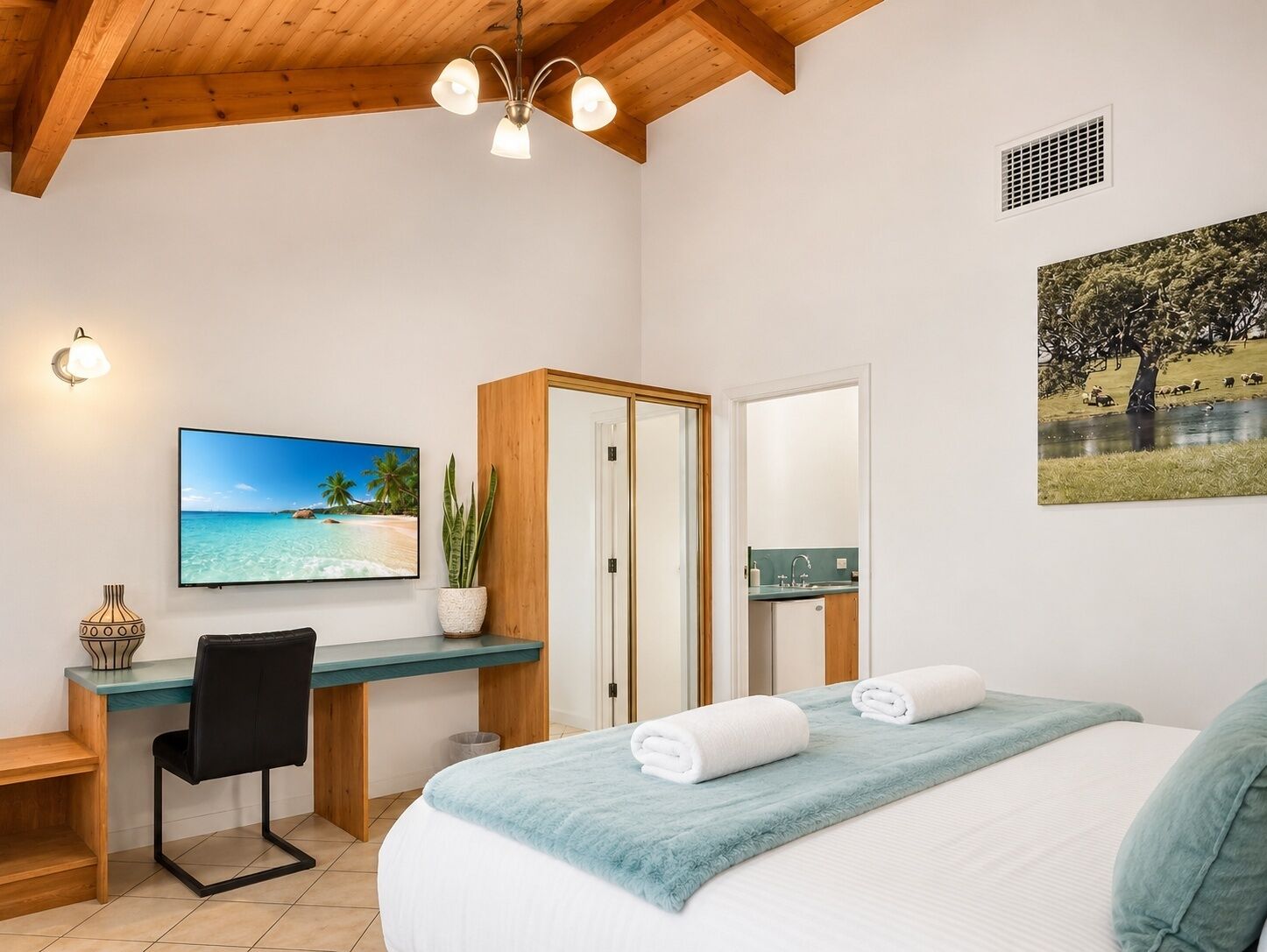 Habitación de hotel con cama, escritorio, silla, televisor, planta y un cuadro.