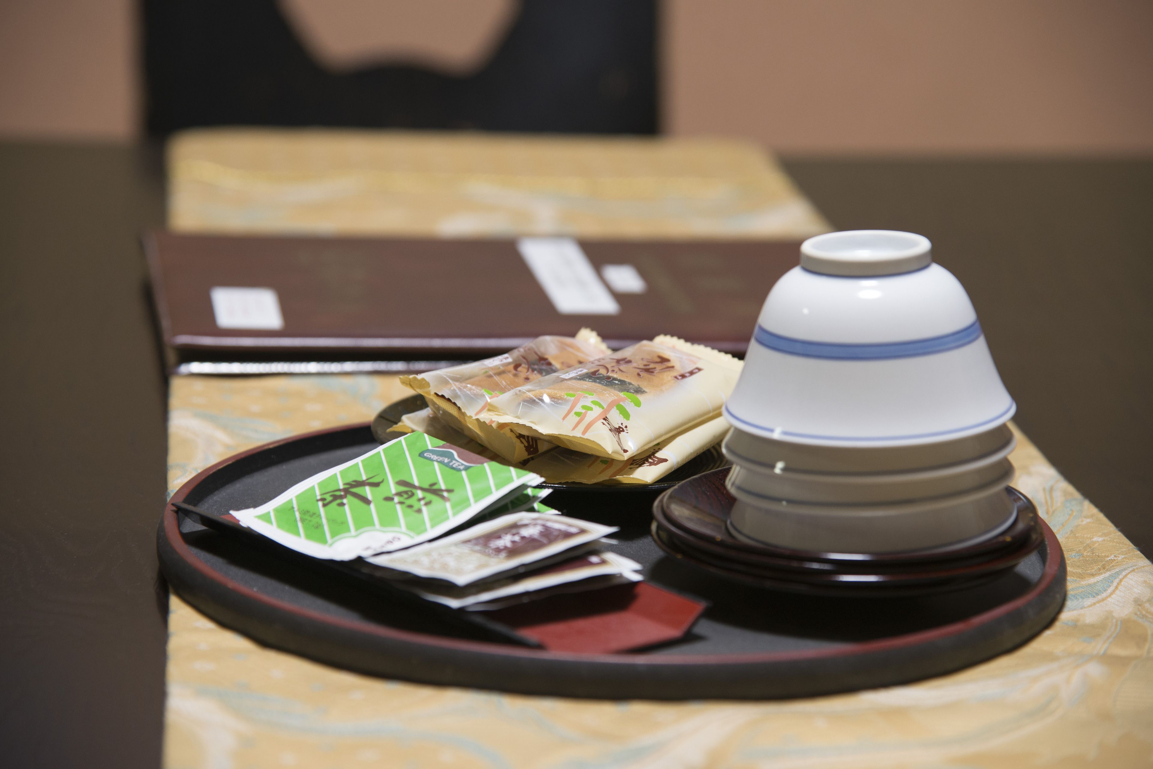 Uma bandeja com um bule, um objeto embrulhado e dois pacotes sobre uma mesa com toalha de mesa estampada.