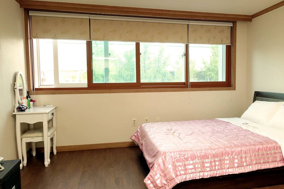 Een slaapkamer met een bed, een nachtkastje, een raam met jaloezieën en uitzicht op groen.