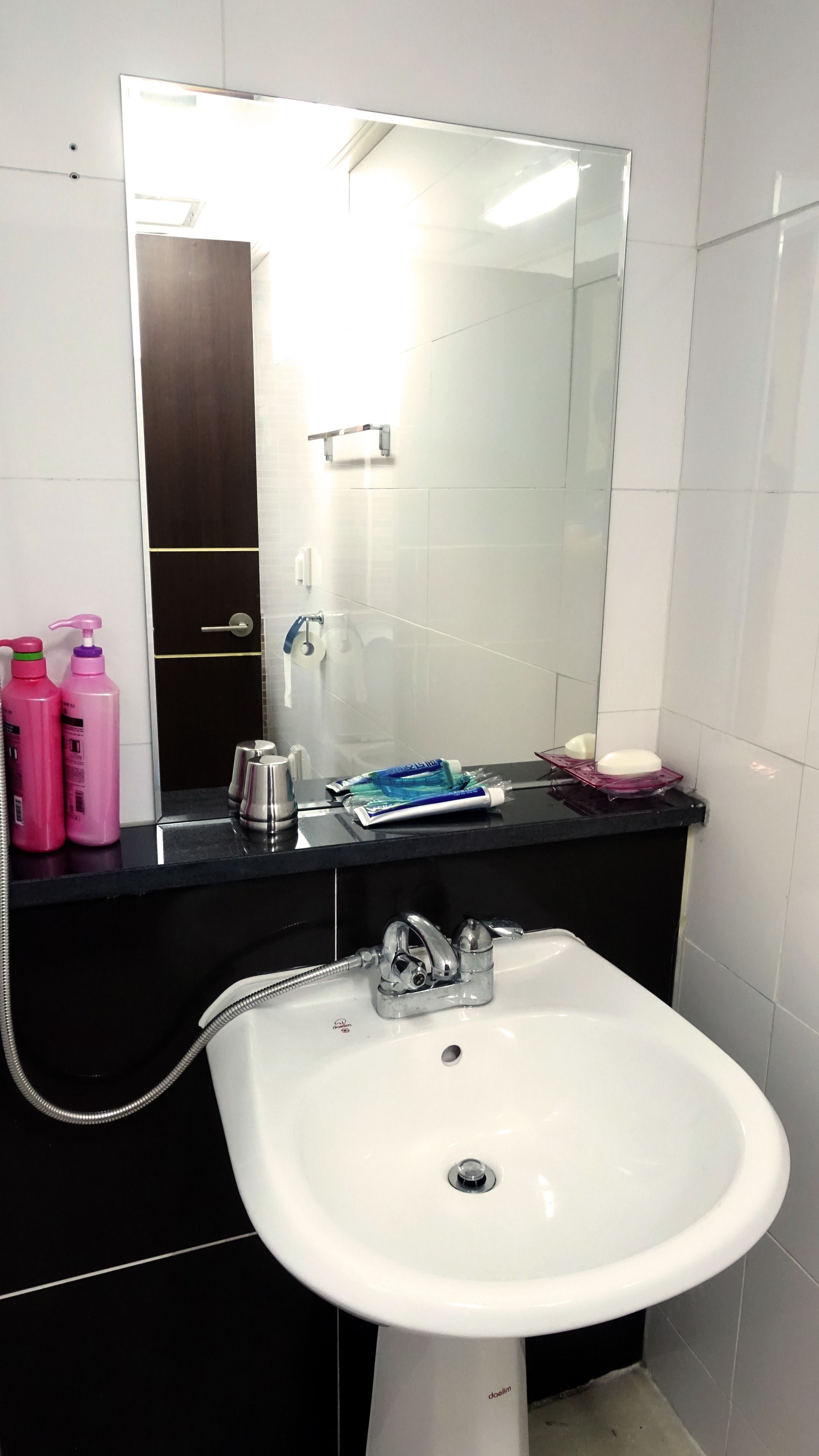 Un baño con un lavamanos blanco, encimera negra y un espejo grande. Hay dos botellas rosas a la izquierda del lavamanos.