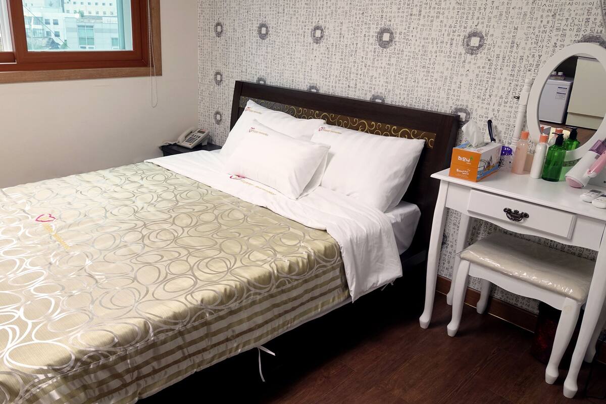 Een net opgemaakt bed met een bedsprei met een patroon, een nachtkastje met toiletartikelen en een raam met uitzicht op gebouwen.