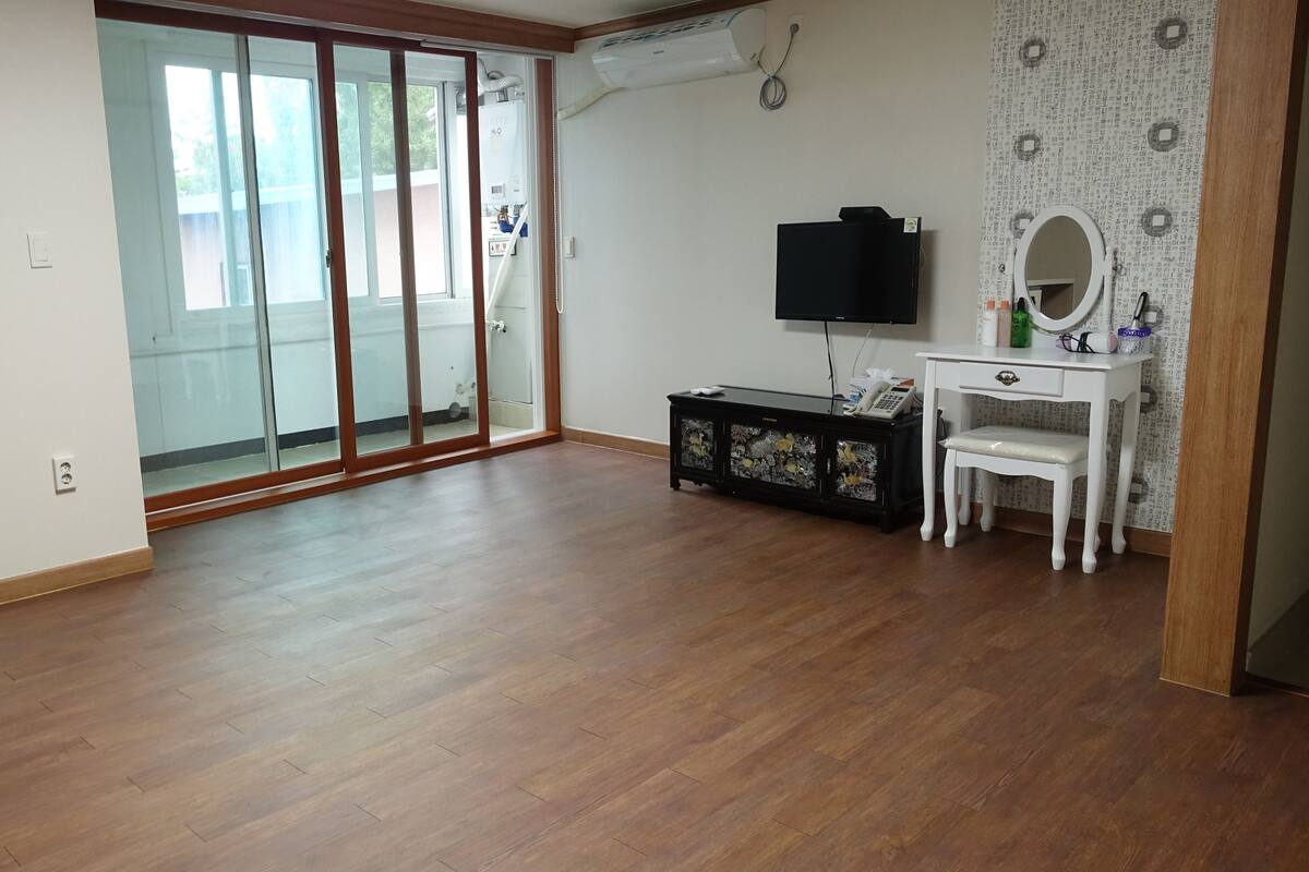 Een kamer met een keukenhoek, een schuifdeur naar een balkon, een wit bureau met een stoel en een zwarte tv-meubel.