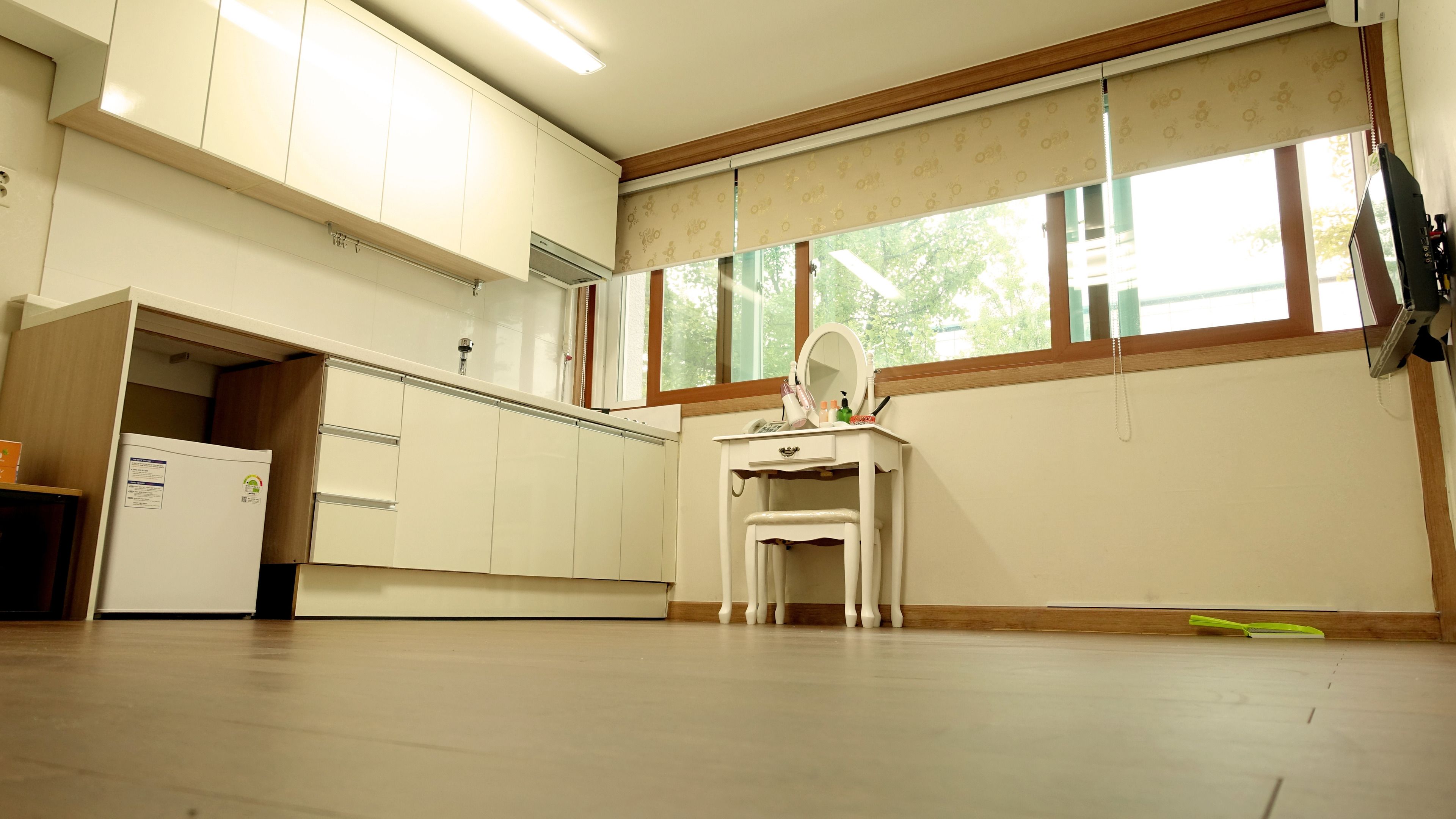 A spacious room with a large window, a white vanity with a mirror, and a refrigerator.
