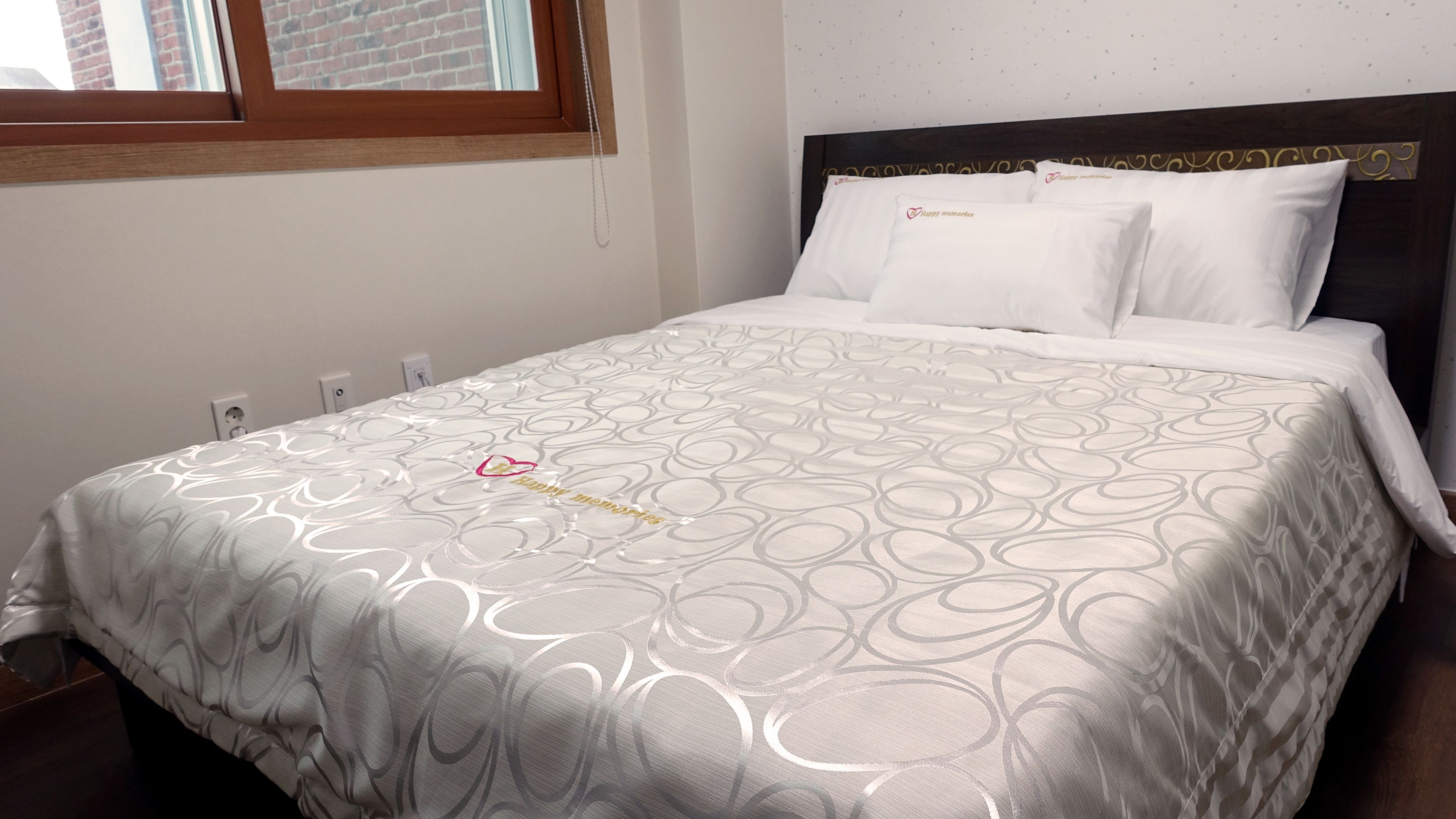A neatly made bed with a patterned bedspread and matching pillows, a wooden headboard, and a window with a view of a brick building.