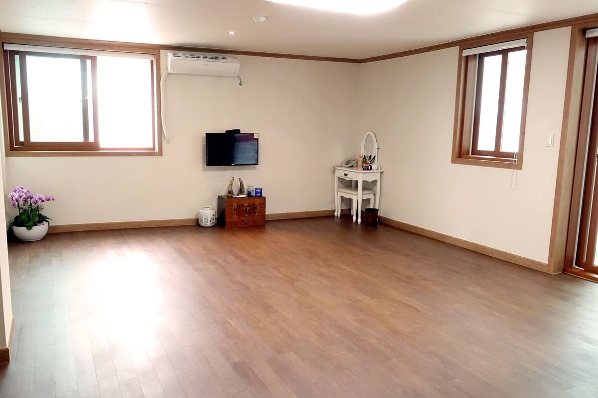 Ruime kamer met houten vloer, een wandtelevisie, een klein wit tafeltje met telefoon en een potplant.
