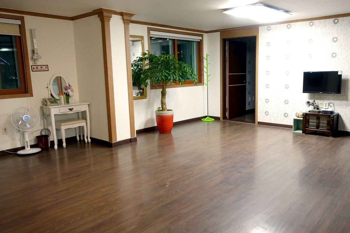 Ruime kamer met houten vloer, een witte kaptafel met spiegel, een potplant, een staande ventilator en een aan de muur bevestigde televisie.