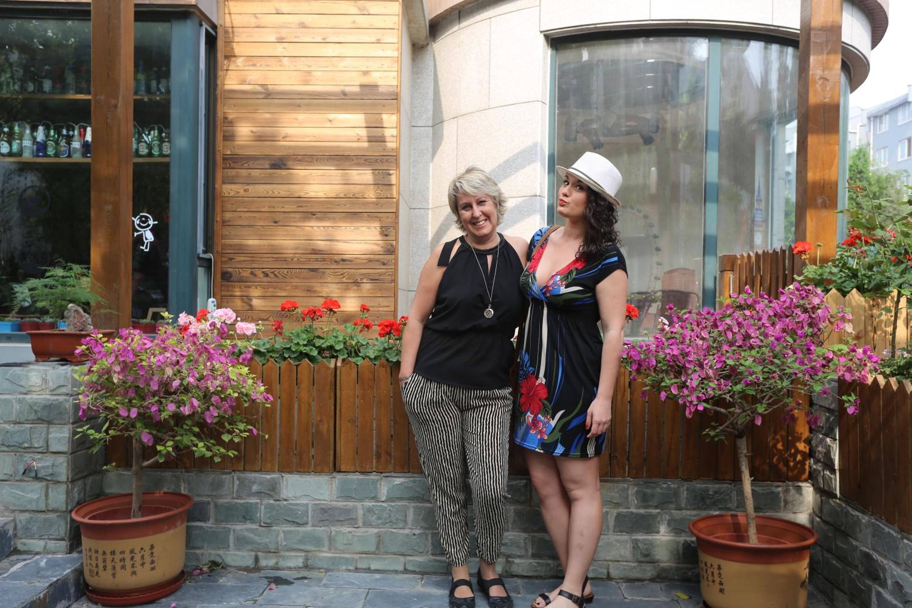 Two people standing in front of a building with wooden paneling and a stone wall. There are potted plants with pink flowers and a wooden fence in the background.