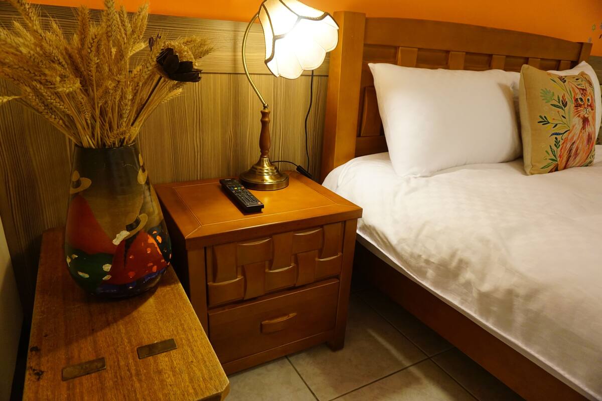 A neatly made bed with white linens, a wooden nightstand with a remote control and a vase of dried flowers, and a night lamp.