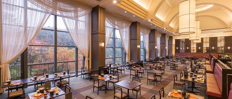 A spacious dining area with multiple tables and chairs, large windows with sheer curtains, and a high ceiling with recessed lighting.