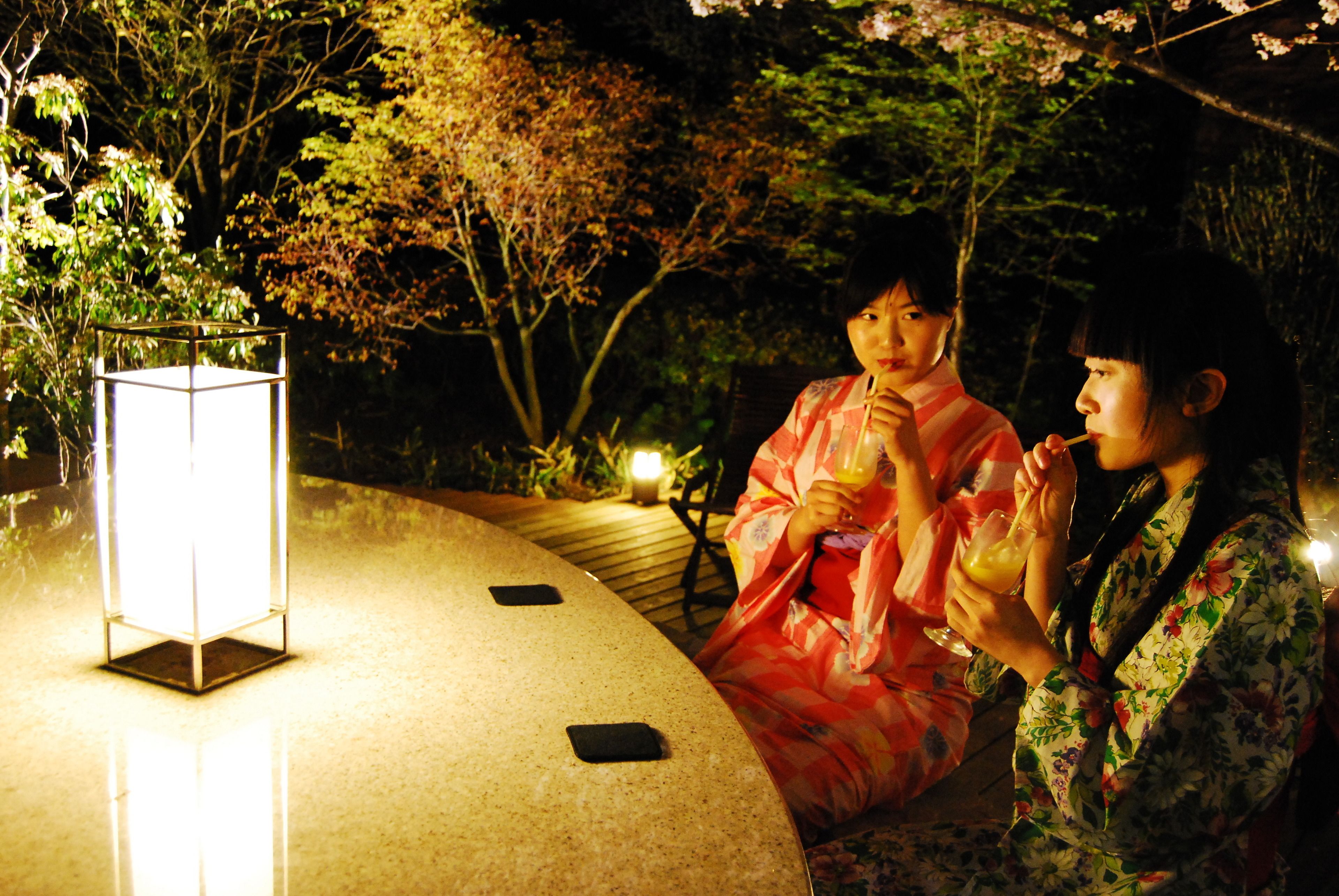 Two individuals in traditional attire sitting at a table with a lit lantern, surrounded by trees and a wooden deck.