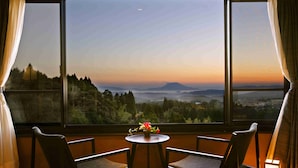 A room with a large window, two chairs, a small table with a flower vase, and a lit table lamp. - Kirishima Kanko Hotel (Kirishima)