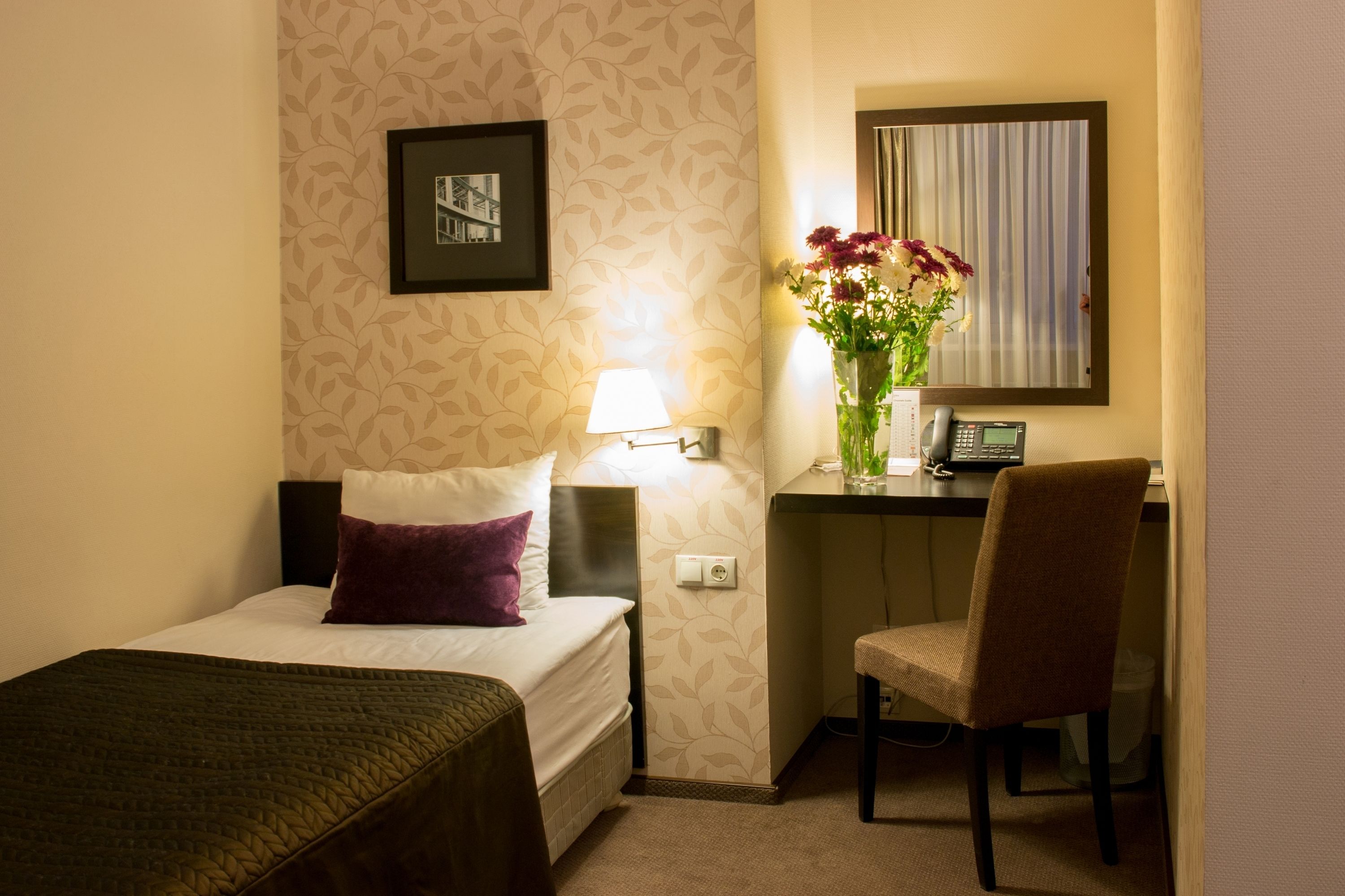 A hotel room with a bed, a desk with a chair, a lamp, a vase with flowers, a phone, and a framed picture on the wall.
