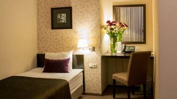 A hotel room with a bed, a desk with a chair, a lamp, a vase with flowers, a phone, and a framed picture on the wall.