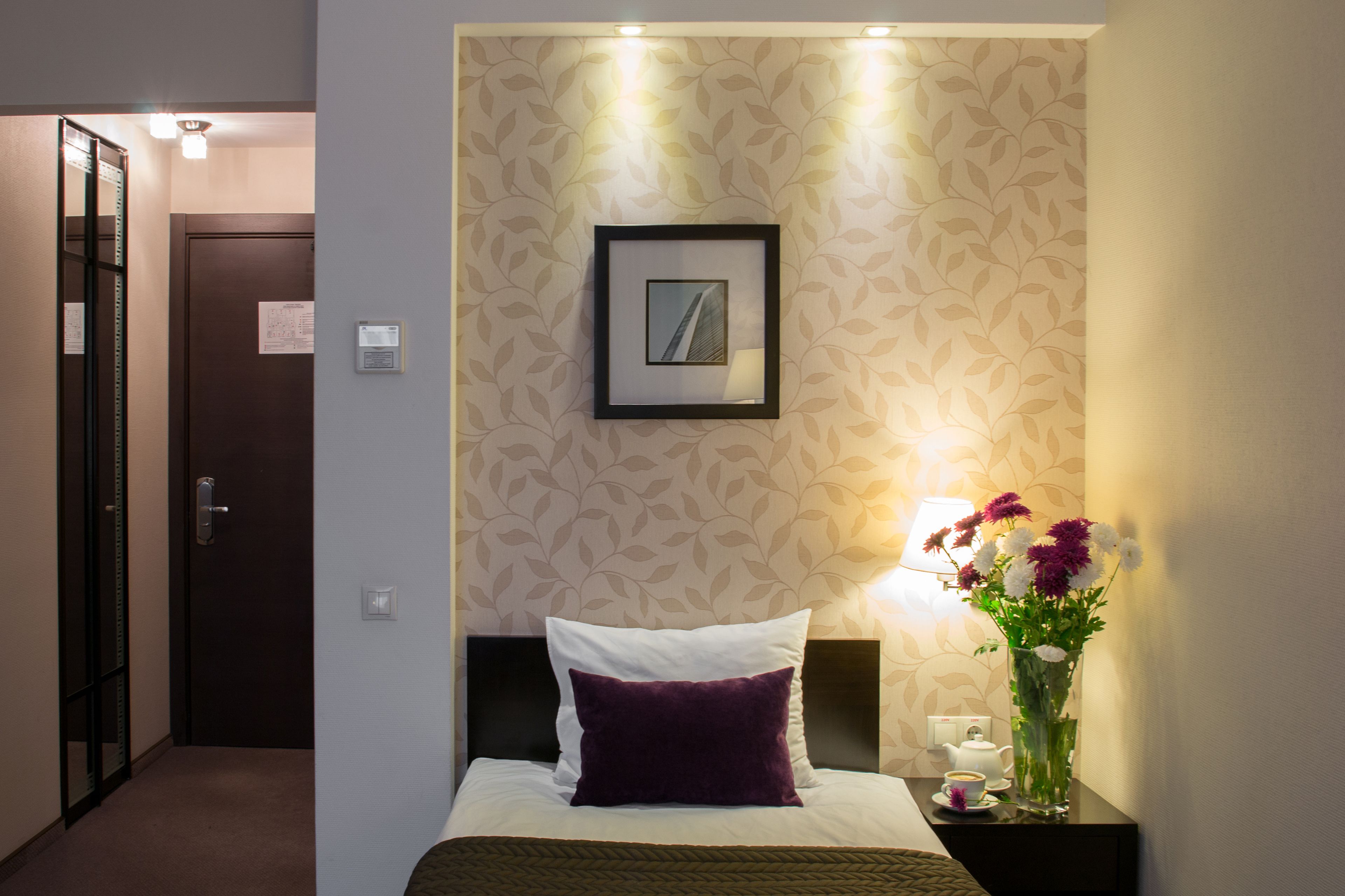Habitación de hotel con cama, mesita de noche con un florero y un cuadro en la pared, además de una puerta al fondo.