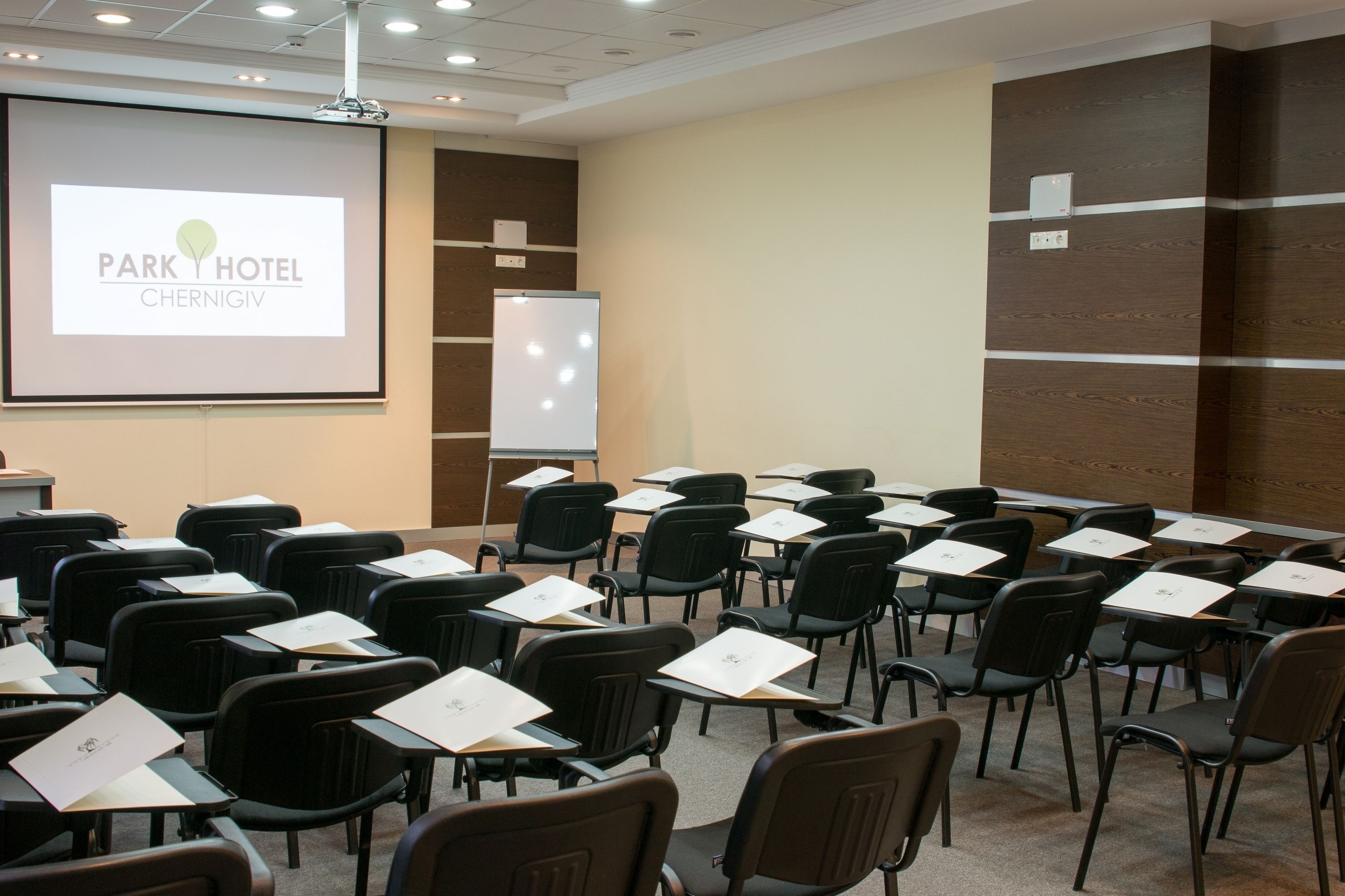 Ein Konferenzraum mit Reihen schwarzer Stühle, einer Projektionsleinwand, die 'Park Hotel Chernihiv' zeigt, und einer Whiteboard-Tafel.