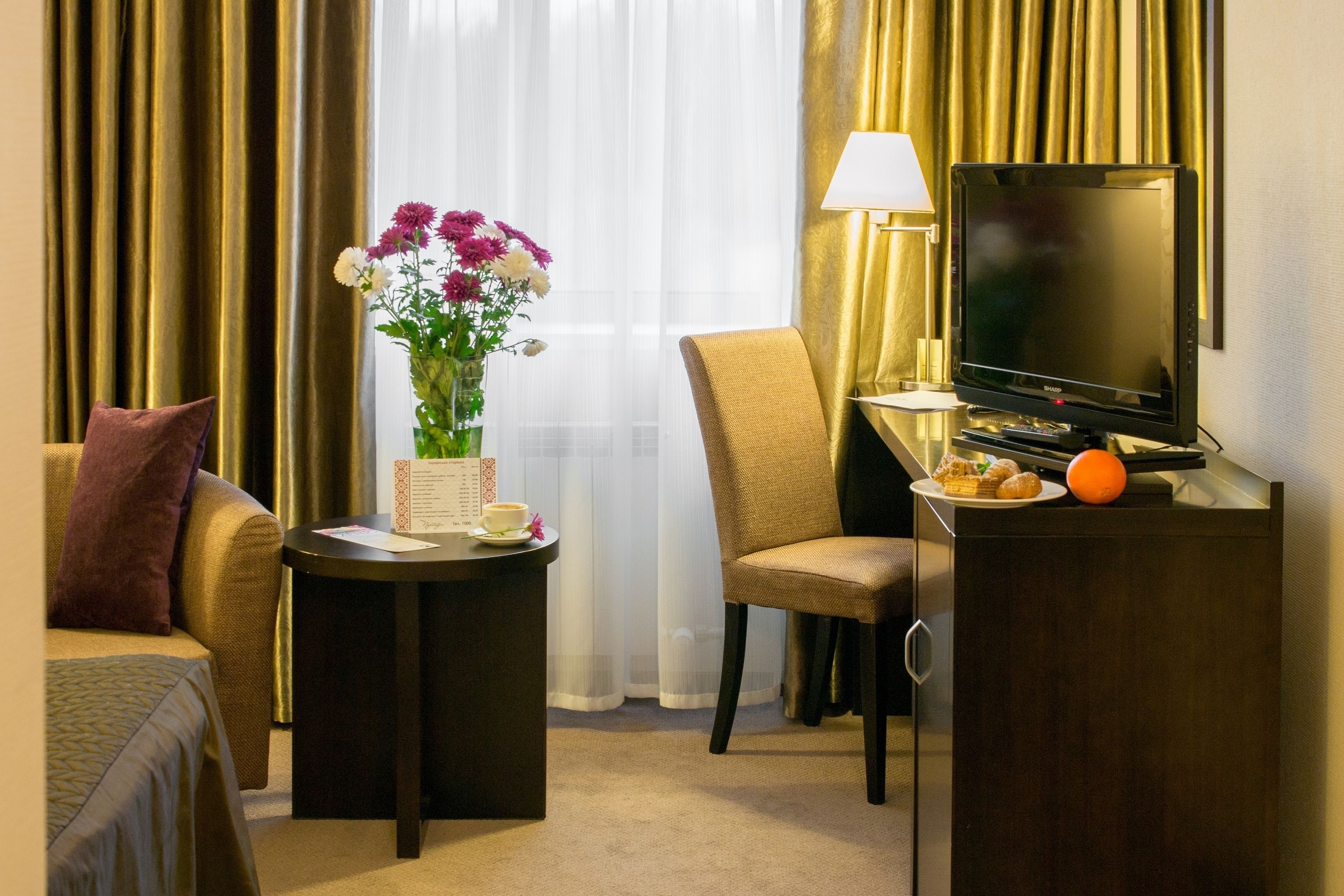 Una camera d'albergo con un letto, una sedia, un tavolino con un vaso di fiori, una lampada, una televisione e un minibar.
