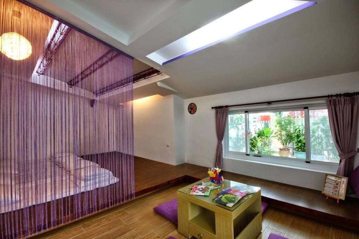 A room with a large window, a wooden floor, a small table with books, and a curtain divider.