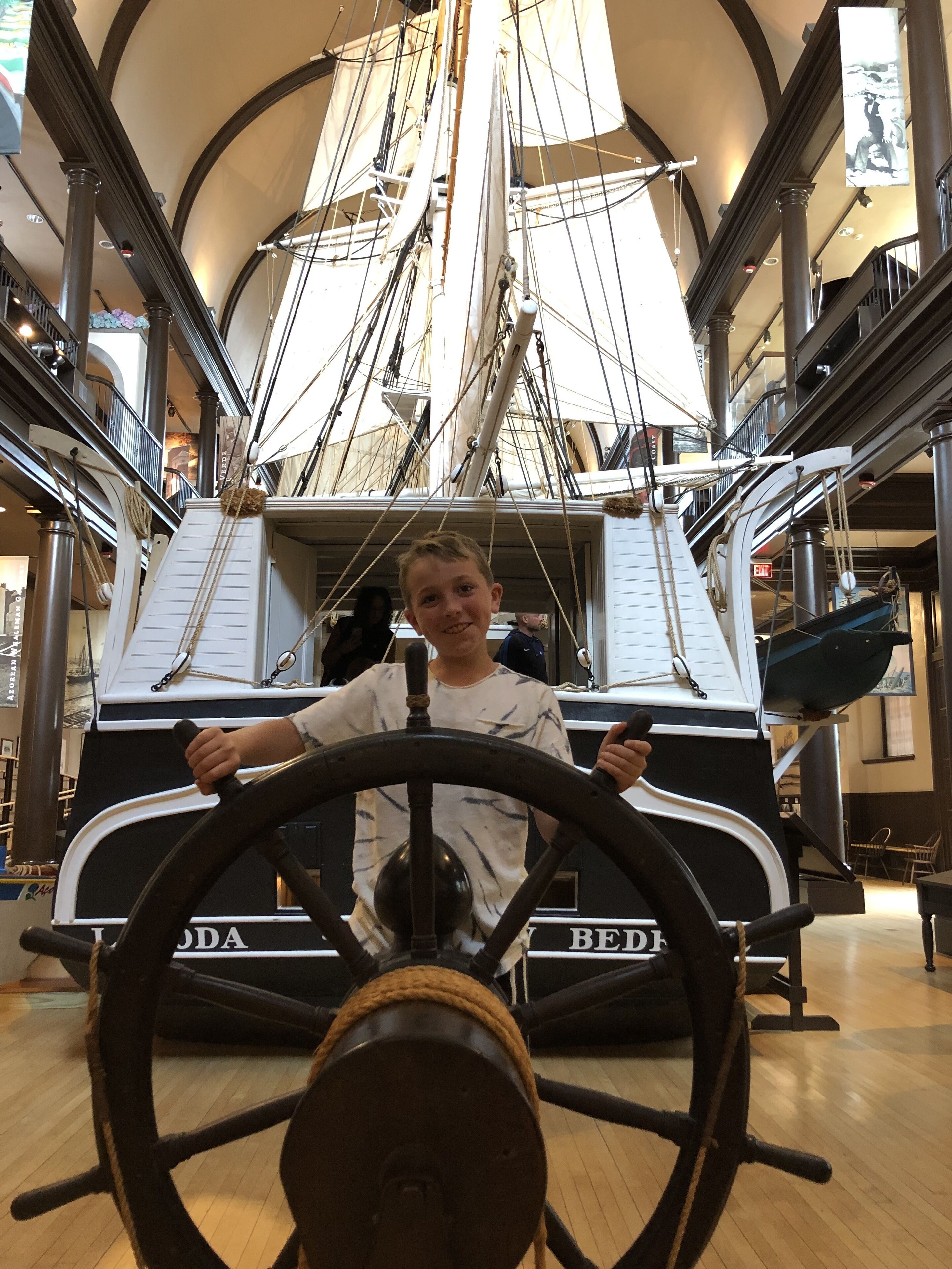 A child in a ship's wheel steering a model ship in a museum.