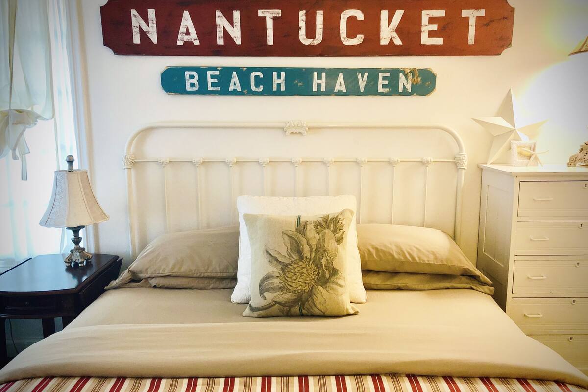 A neatly made bed with two pillows, a striped bedspread, and a wall sign reading "NANTUCKET BEACH HAVEN".