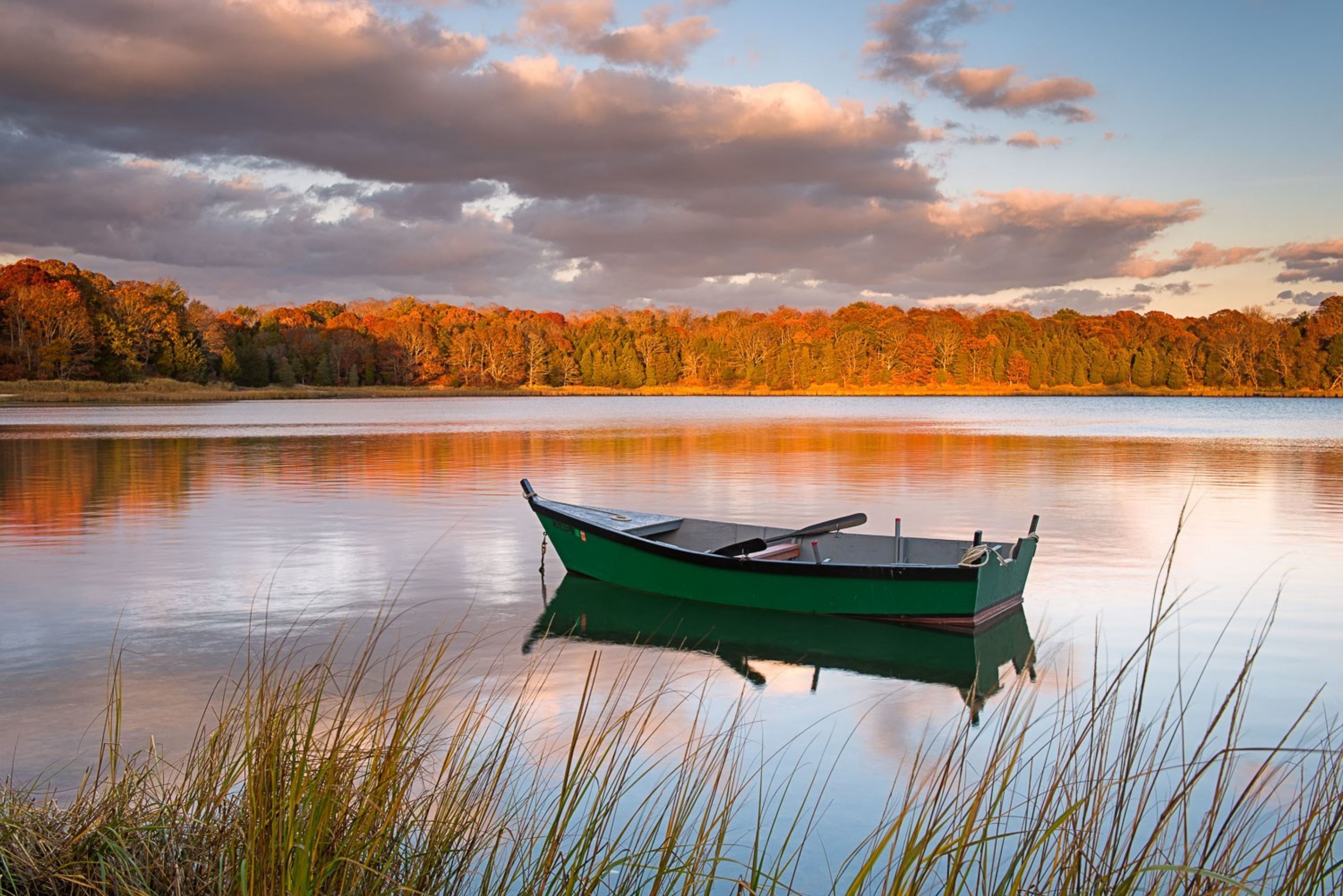 A calm lake with a green boat anchored near the shore, surrounded by autumn foliage.