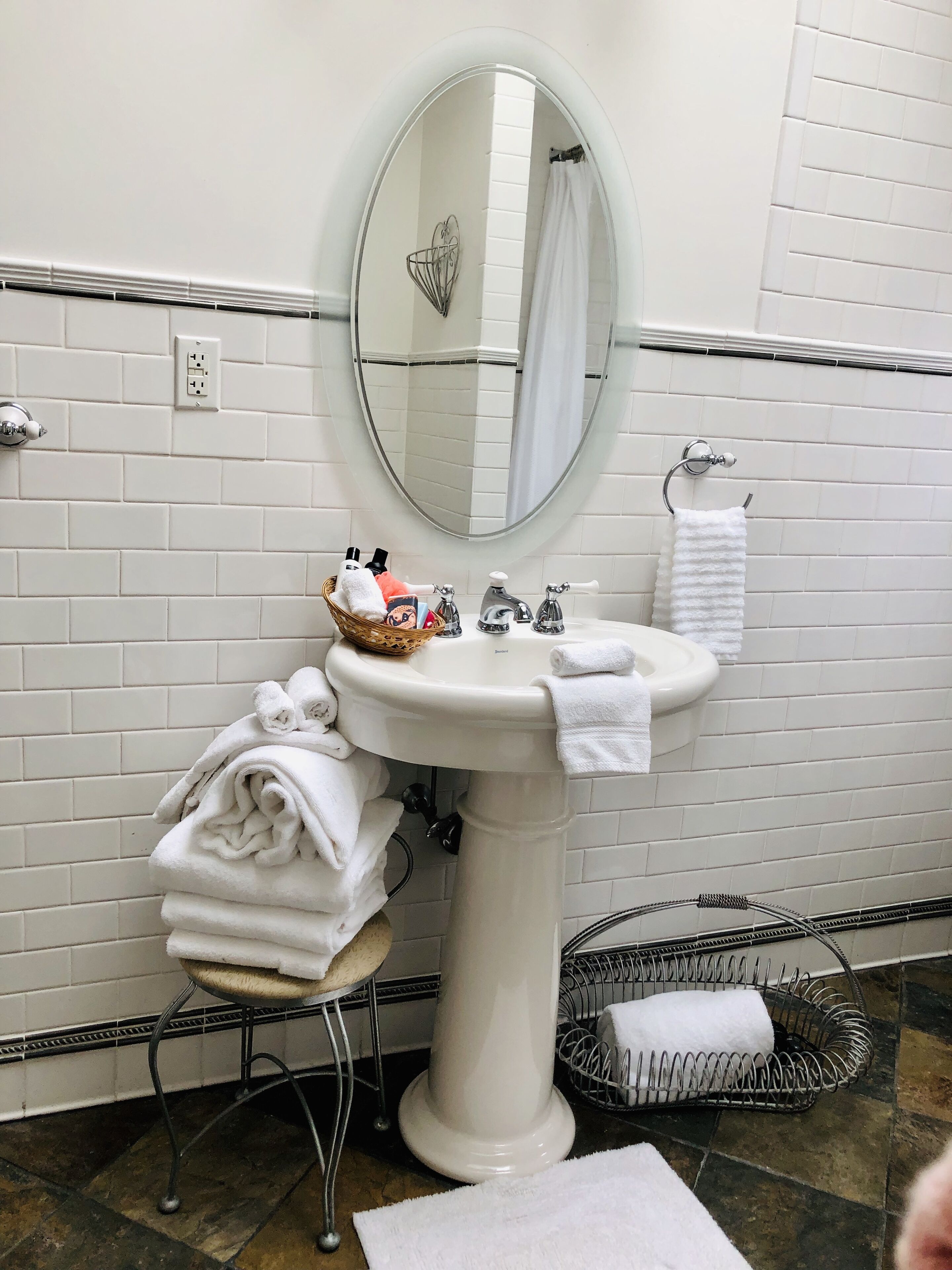 Ein Badezimmer mit weiß geflieste Wand, einem Standwaschbecken, einem runden Spiegel, einem Handtuchhalter und einem Metallhocker mit Handtüchern.