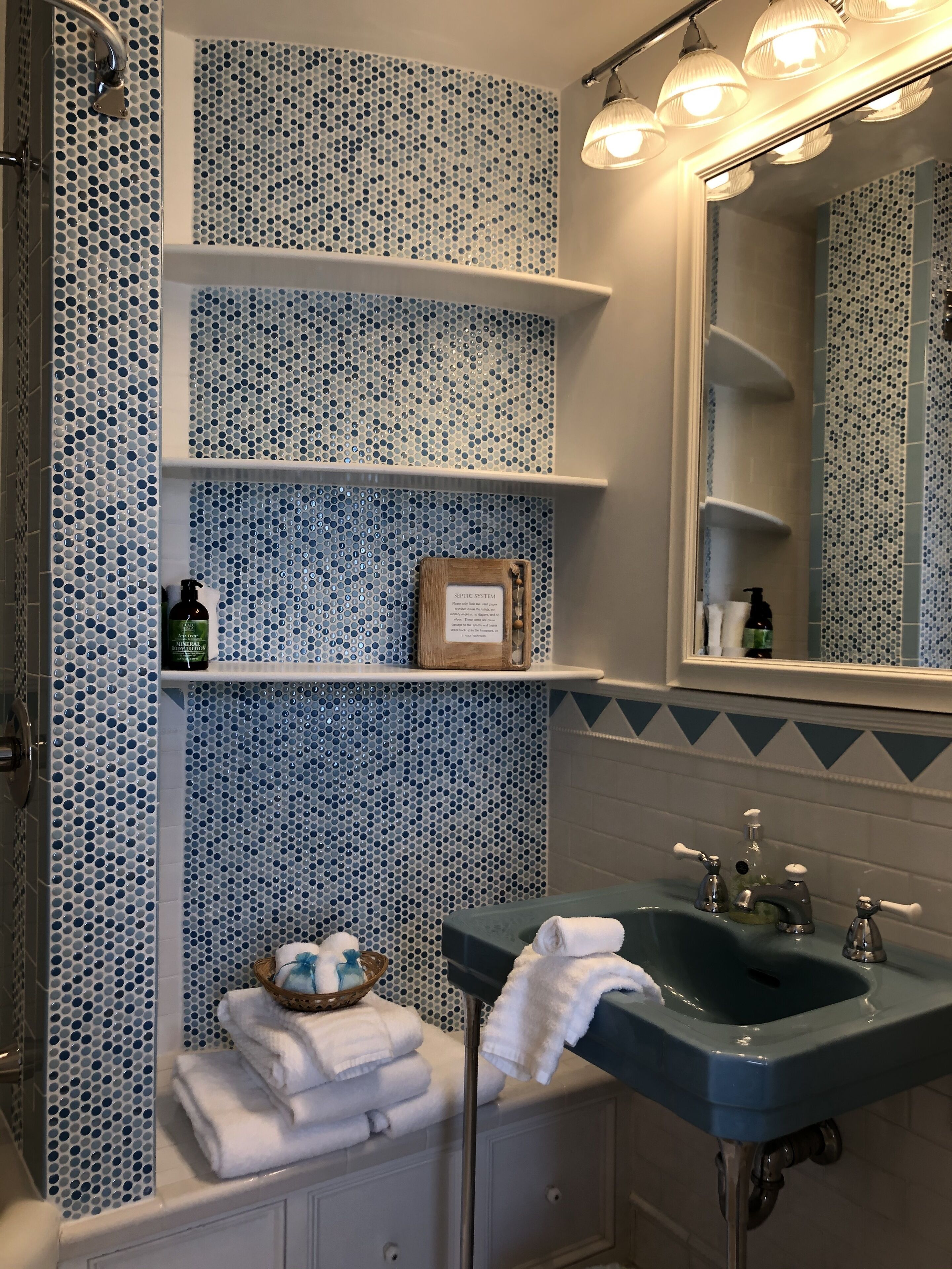A bathroom with a blue and white mosaic tile wall, a sink with two faucets, and a mirror above the sink. Shelves are mounted on the wall, holding toiletries and a framed certificate.