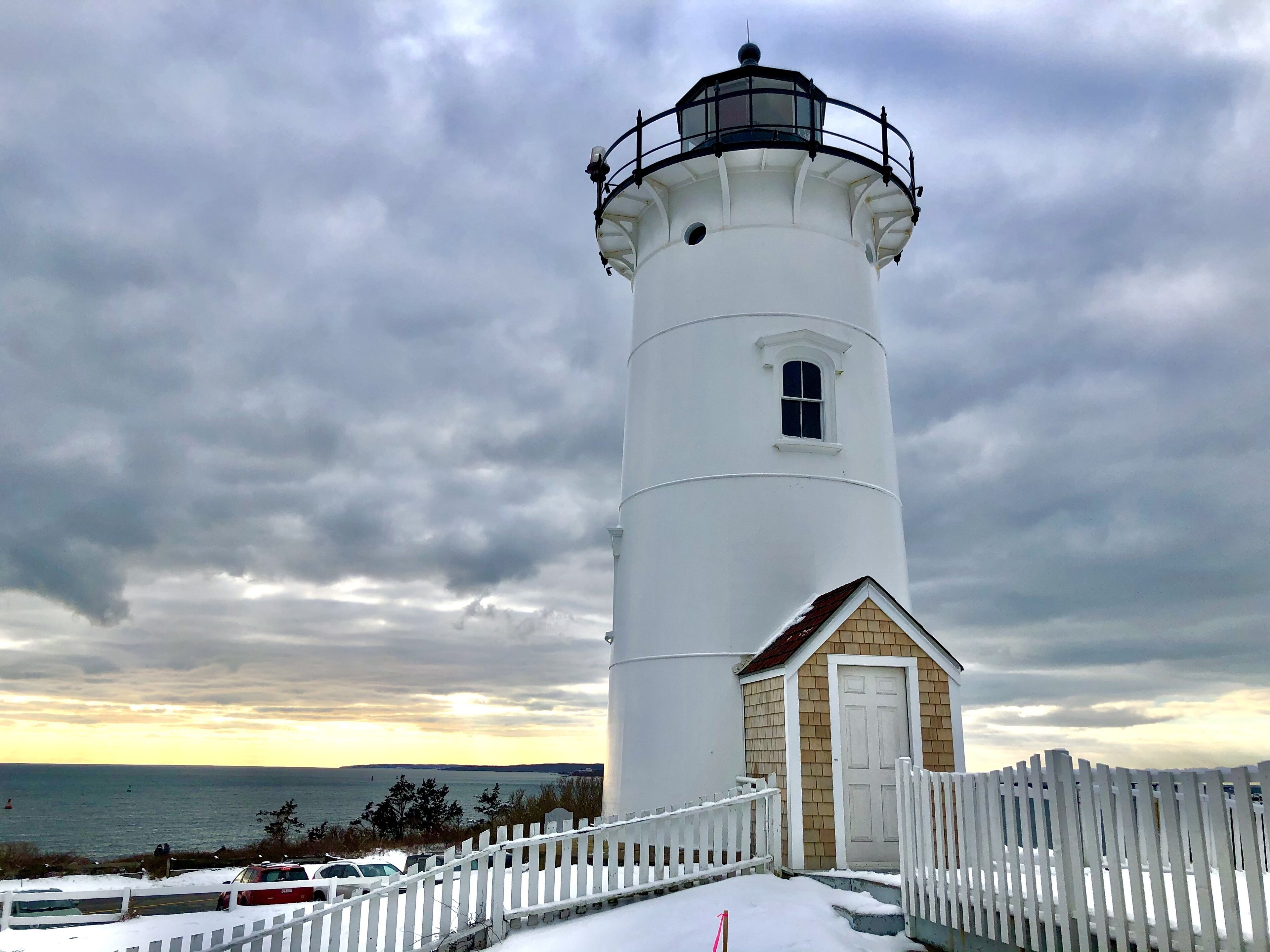 A white lighthouse with a small attached building, a white picket fence, and a view of the sea.