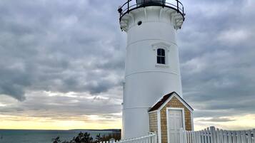A white lighthouse with a small attached building, a white picket fence, and a view of the sea.
