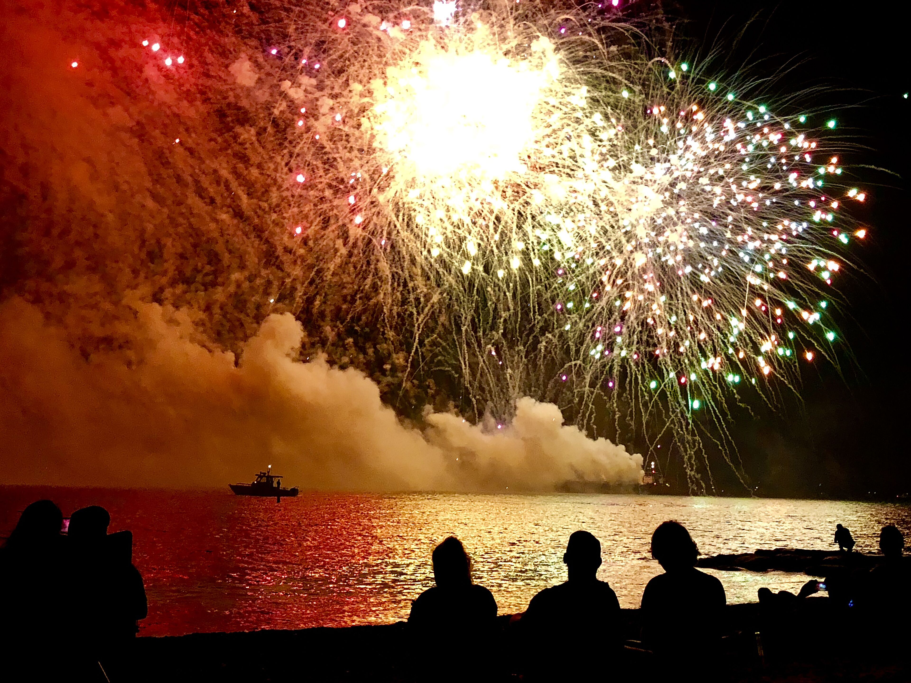A nighttime scene with fireworks over a body of water, silhouettes of people watching, and a boat in the distance.