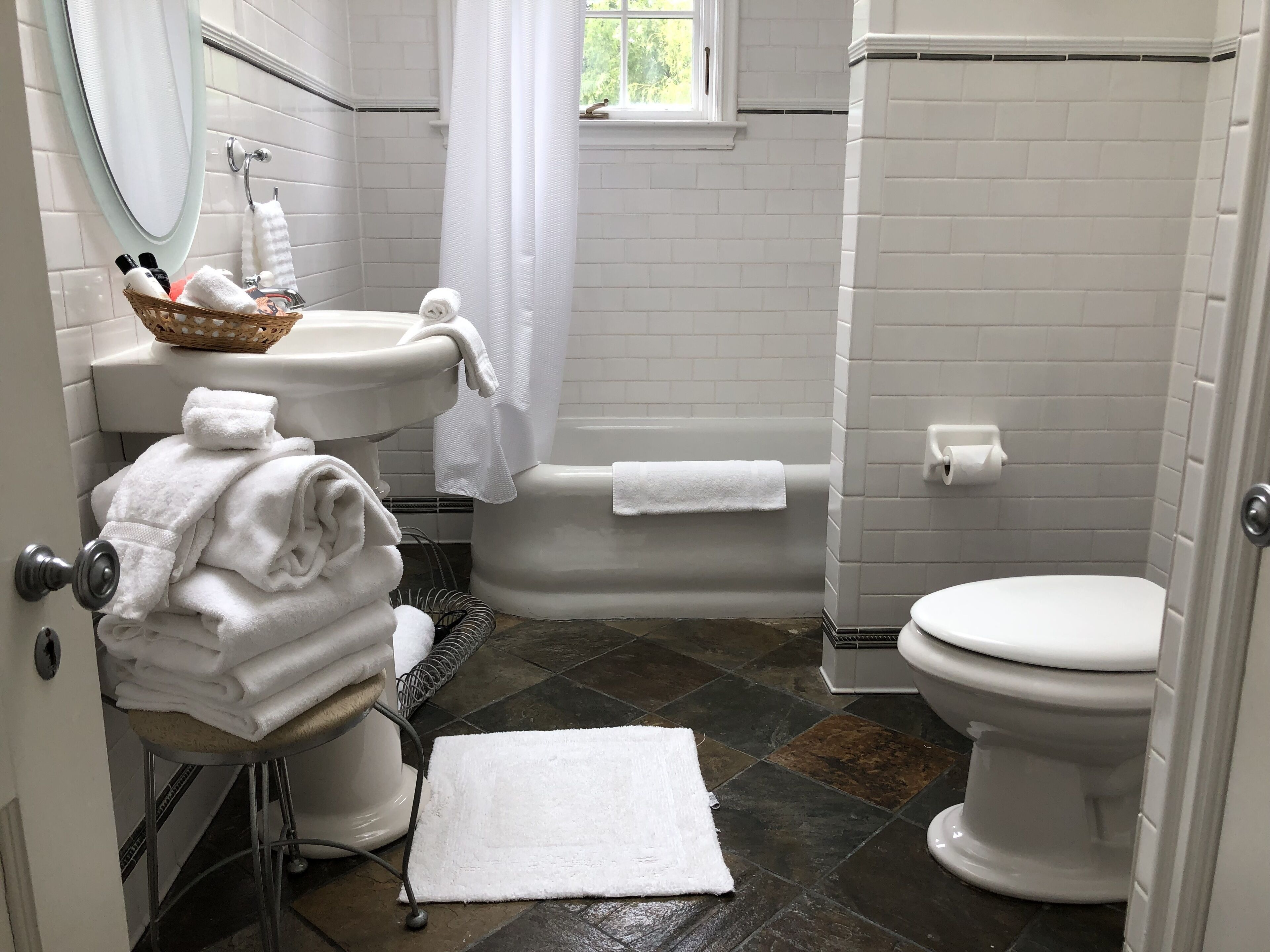 A bathroom with a sink, toilet, and bathtub. There are towels stacked on a chair and a basket of toiletries on the sink.