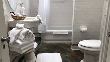 A bathroom with a sink, toilet, and bathtub. There are towels stacked on a chair and a basket of toiletries on the sink.