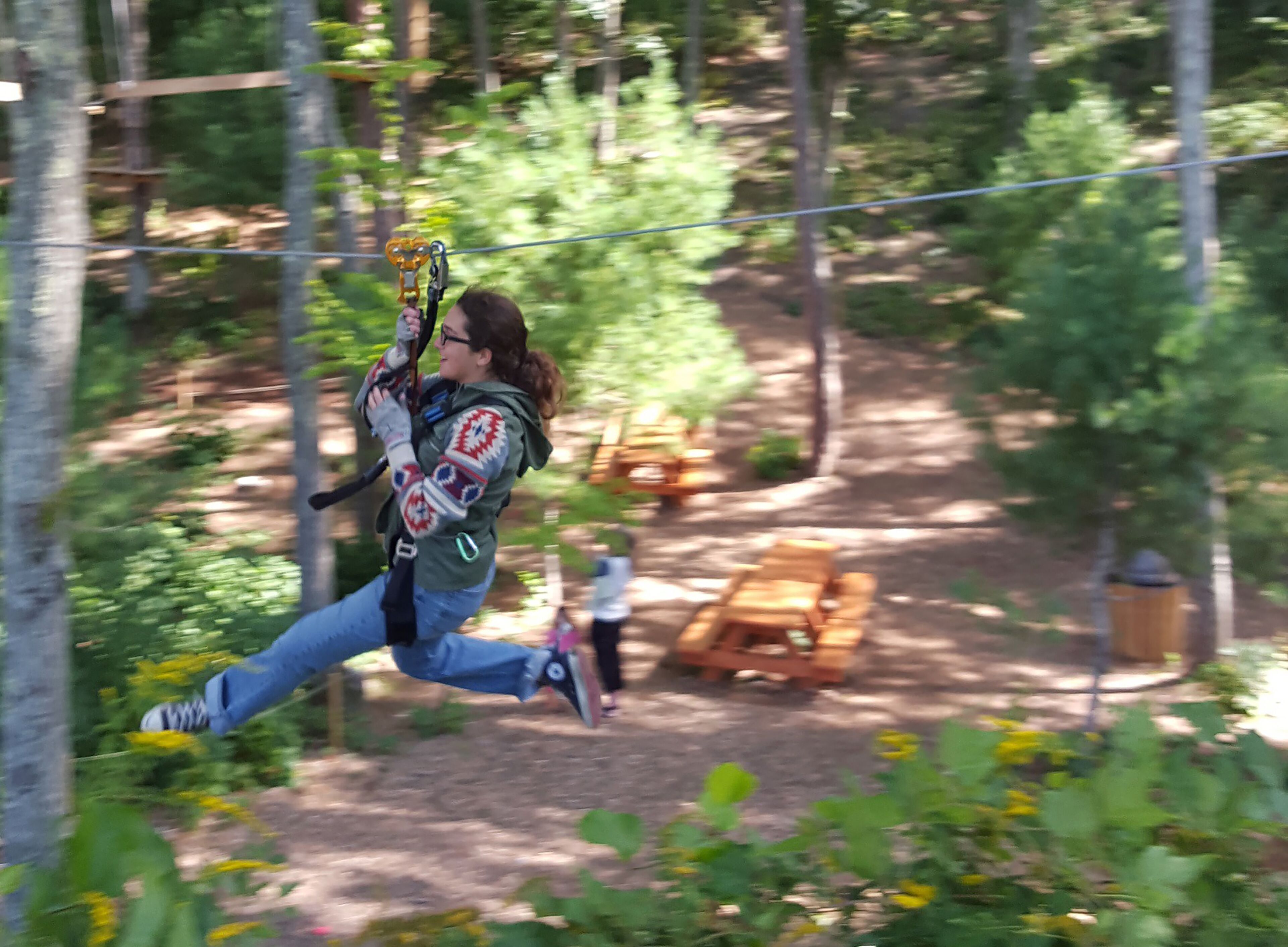 A person zip-lining through a wooded area with picnic tables and a trash can in the background.