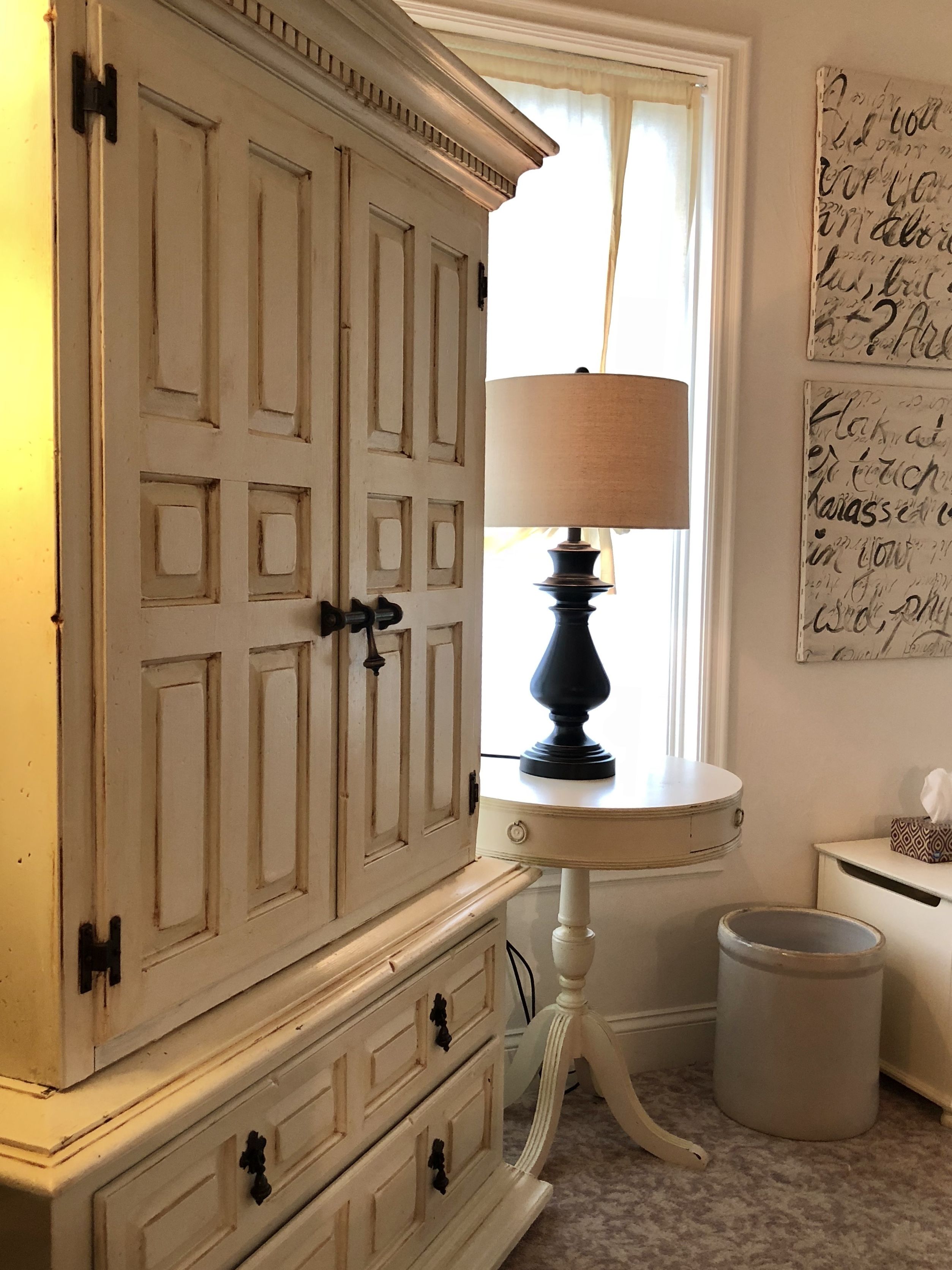 A room with a large white cabinet with multiple drawers, a small white table with a lamp, and a trash can.