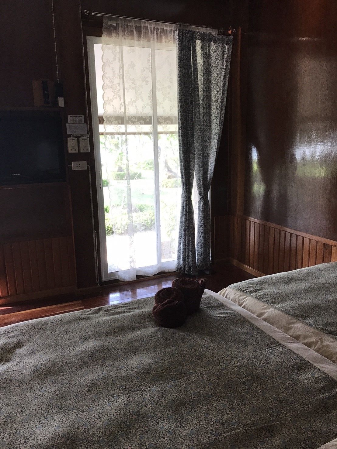 A room with a wooden floor, a bed with a grey and white bedspread, a pair of slippers on the floor, and a window with patterned curtains.