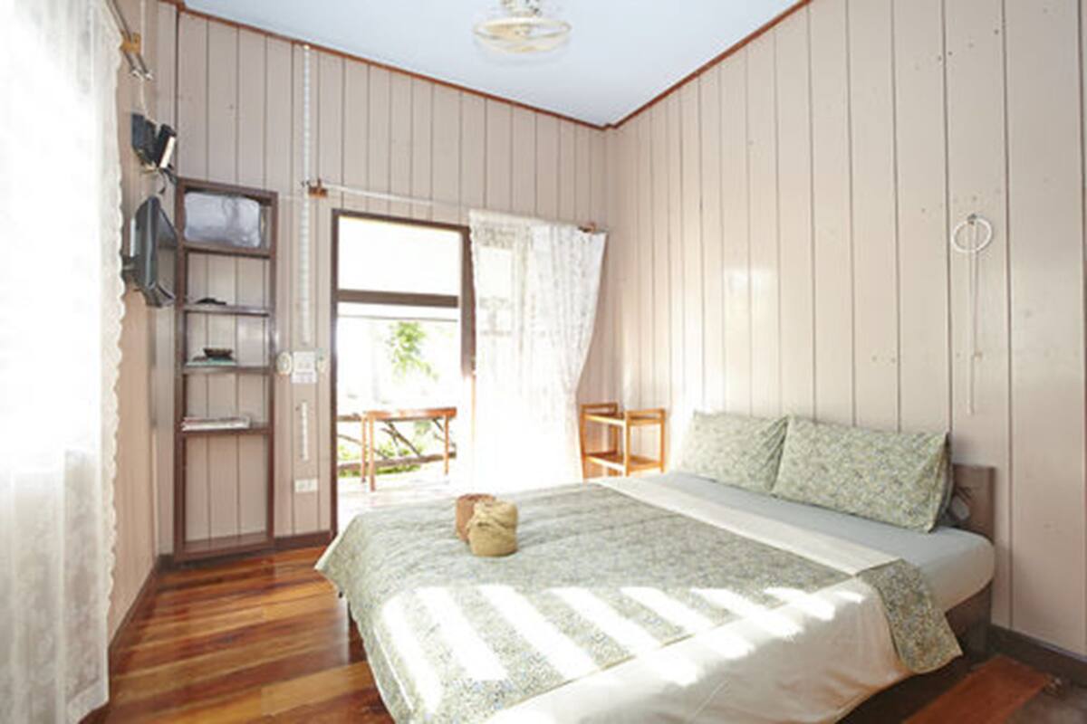 Ein Schlafzimmer mit Holzboden, einem Bett mit floralem Überwurf, einem Fenster mit Vorhängen und einem kleinen Tisch mit Stuhl.