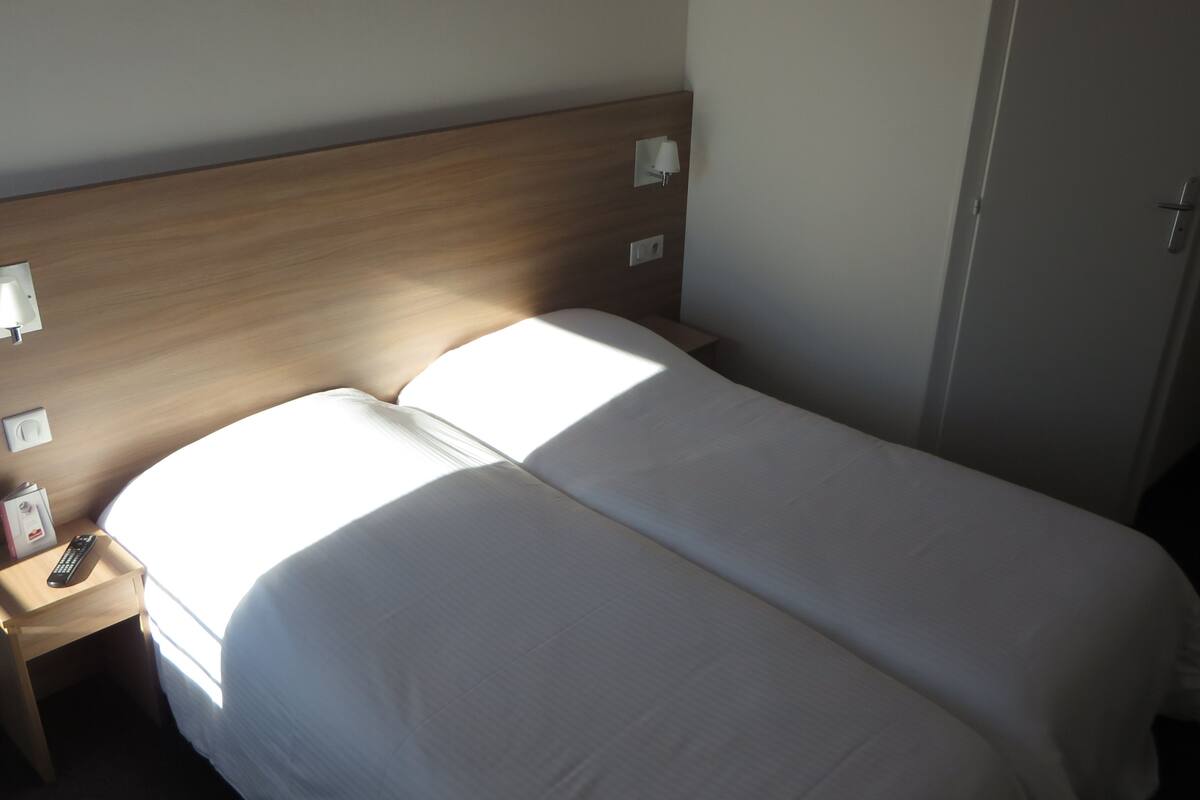 Ein Hotelzimmer mit zwei Einzelbetten, einem Nachttisch mit Lampe, einer Fernbedienung und einer Tür.